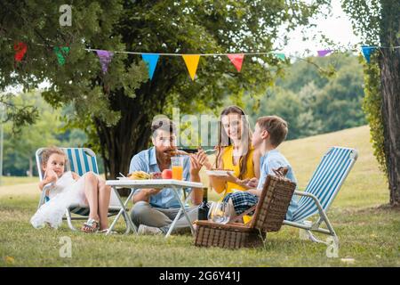 Dedicated young parents of two children listening to their funny son talking while eating together during family picnic in a summer day Stock Photo