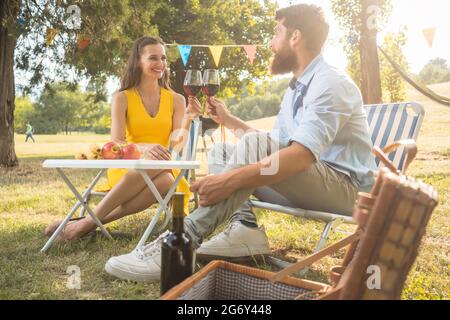 Young and beautiful couple in love toasting with red wine while sitting down on folding chairs during romantic picnic in the countryside Stock Photo