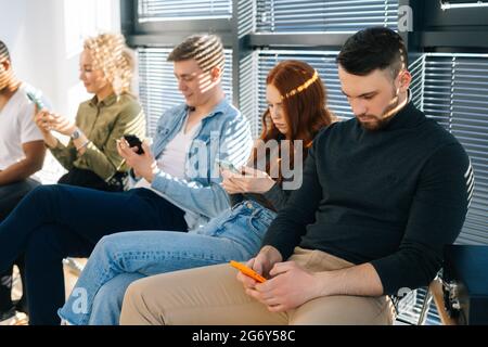 Side view of group five young diverse ethnicity candidates using mobile phones while waiting job interview in modern office lobby. Stock Photo