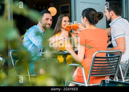 Cheerful young best friends toasting with a refreshing delicious summer drink while sitting together at table at a trendy restaurant outdoors Stock Photo