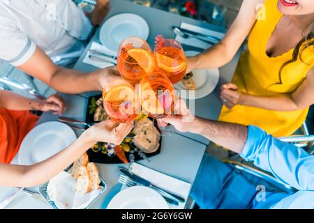 High-angle view of the hands of four friends holding stemmed wine glasses with a delicious orange summer drink, while toasting during lunch outdoors a Stock Photo