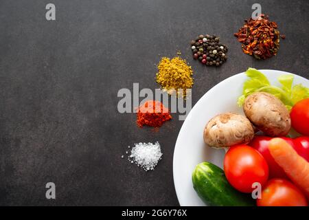 A white plate with fresh vegetables: mushrooms, tomatoes, carrots and herbs, around which various spices are scattered in piles. On a black background Stock Photo