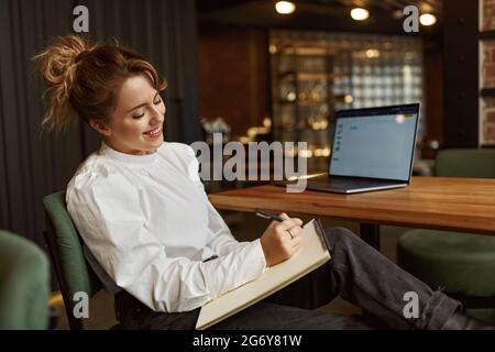 Smiling woman taking notes while working on laptop Stock Photo