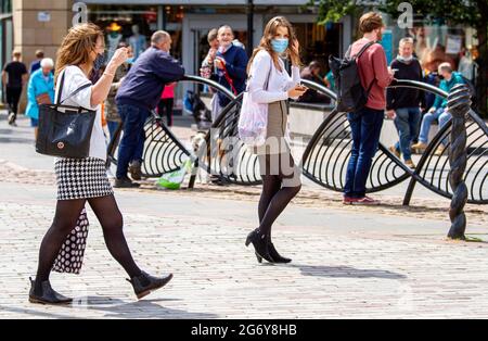 Dundee, Tayside, Scotland, UK. July, 2021. UK Weather: Humid day with a warm breeze and sunny intervals across North East Scotland with temperatures reaching 18°C. Two fashionable women together outside wearing facemasks holding their I Phones whilst enjoying the warm sunshine in Dundee city centre. Credit: Dundee Photographics/Alamy Live News