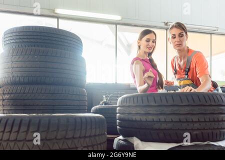Helpful experienced female auto mechanic checking the identification number of a tire for a customer in an automobile repair shop with various tires f Stock Photo
