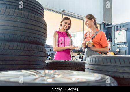 Helpful experienced female auto mechanic checking the identification number of a tire for a customer in an automobile repair shop with various tires f Stock Photo