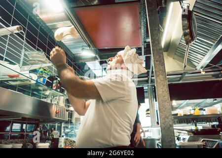 Low-angle side view of an experienced cook tossing dough while preparing pizza in the kitchen of a modern Italian restaurant Stock Photo