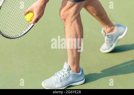 Low section of a professional player wearing gray sport shoes while holding the ball and the tennis racket during match on green surface Stock Photo