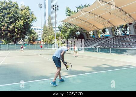 Rear view of a man ready to serve while standing behind the baseline of a professional tennis court during doubles match Stock Photo