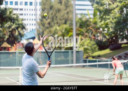 Rear view of a man ready to serve while standing behind the baseline of a professional tennis court during doubles match Stock Photo