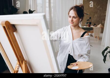 Young creative woman in earphones painting on easel Stock Photo