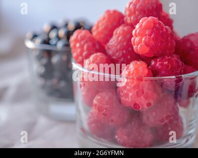 Closeup of fresh ripe raspberries in a low transparent glass and a glass of currant berries at the blurry background. Stock Photo