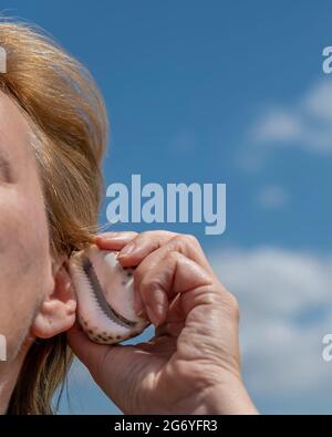 A blonde woman brought a shell to her ear to hear the sound of the sea Stock Photo
