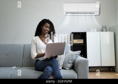 Young Happy Woman Sitting On Couch Using Air Conditioner At Home Stock Photo