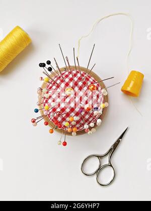 Round pin cushion with lot of pins, yellow thimble, spool of thread and scissors. Sewing accessories flat lay on white background Stock Photo