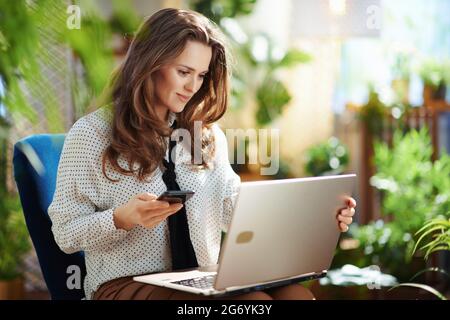 Green Home. smiling elegant woman with long wavy hair with smartphone and laptop sitting in a blue armchair at modern home in sunny day. Stock Photo