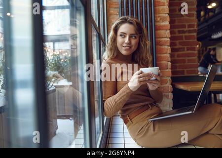 Pleasant woman drinking coffee and using laptop at cafe Stock Photo