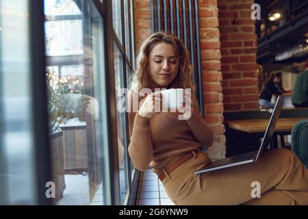 Woman using laptop and drinking coffee near window Stock Photo