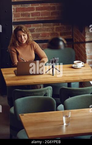 Smiling woman using laptop and smartphone on tripod at cafe Stock Photo