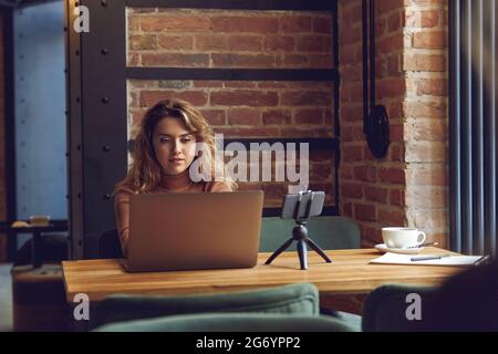 Woman working on laptop at table with smartphone on tripod Stock Photo
