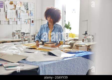 Stylish African-American lady draws pattern on fabric using tailors chalk in modern workshop Stock Photo