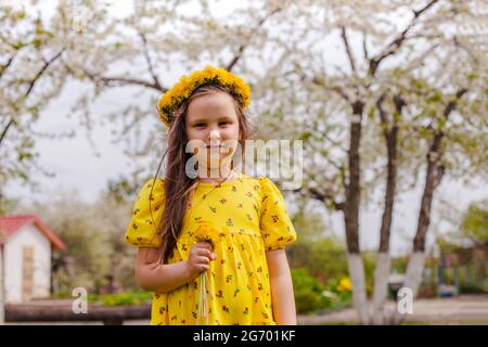 lifestyle portrait of a beautiful girl in a yellow dress and a wreath of yellow dandelions on her head, with spring blooming cherry trees in the Stock Photo