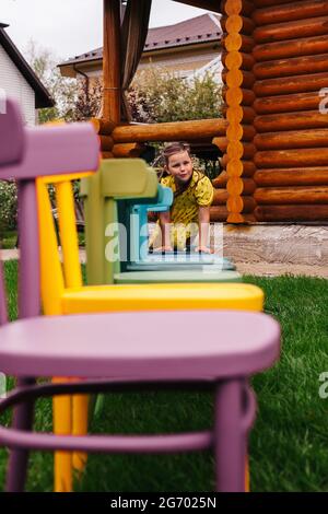 a cheerful girl crawls along a row of colorful wooden chairs, a girl has a good time in the backyard Stock Photo