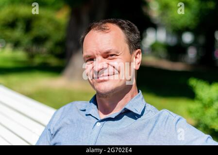 A Caucasian man of forty years on the street in the summer sits in a park on a bench and smiles. Stock Photo