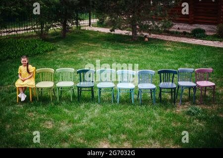 top view of a girl sitting on the edge in a row of colorful wooden chairs and waiting for guests for a children's birthday in the garden on a sunny Stock Photo