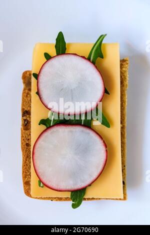 Sandwich made from rye bread, hard cheese, radish, arugula, served on a white plate against a dark wooden background. Symmetry. Copy space. Flat lay, Stock Photo