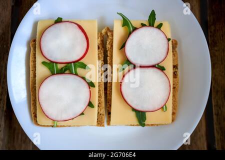 Sandwich made from rye bread, hard cheese, radish, arugula, served on a white plate against a dark wooden background. Symmetry. Copy space. Flat lay, Stock Photo