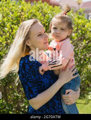 A young beautiful mother looks at her daughter toddler. A girl in her arms. A sunny day. The happiness of motherhood Stock Photo