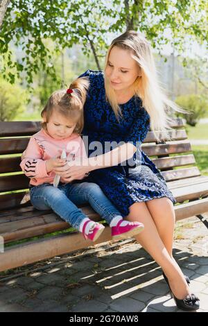 A young beautiful woman and a little toddler girl are sitting on a park bench and looking at a toy. Mother and daughter. A sunny summer day Stock Photo