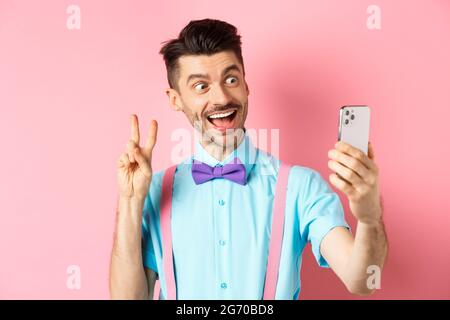 Technology concept. Funny man with moustache and bow-tie taking selfie on smartphone, showing peace sign and smiling at mobile camera, pink background Stock Photo