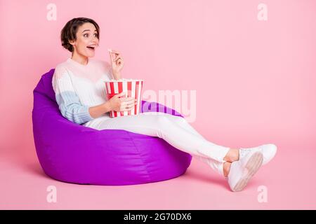 Full length photo portrait of excited girl eating popcorn sitting in violet beanbag chair isolated on pastel pink colored background Stock Photo