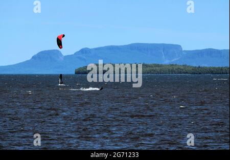 In the Bay at Mission Marsh two men are windsurfing and parasailing in Lake Superior, with the Sleeping Giant and one Welcome Island in the background.