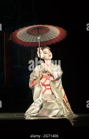 Japanese woman in traditional kimono with umbrella sitting on the knees in darkness, lighten by the moonlignt. Traditional Japanese performance. Stock Photo