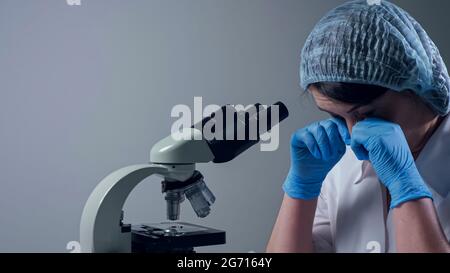 Woman employee in cap and white uniform tired of looking through contemporary microscope closes eyes at workplace in clinic laboratory closeup Stock Photo