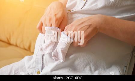 Unrecognizable pregnant woman moves fingers over belly putting them on little socks. Stock Photo