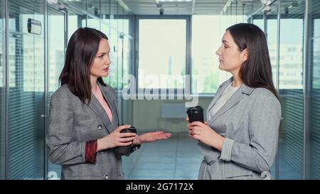 Brunette ladies successful company managers in grey jackets discuss project and give high five with satisfaction standing in corridor against large windows Stock Photo