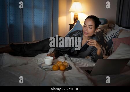 woman in robe lying on bed at home Awake late at night trying work on laptop computer overtime late night. Stock Photo