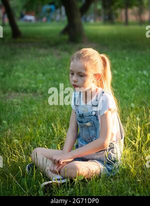 Little sad girl sitting in the park on the grass Stock Photo