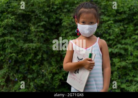 Little asian girl wearing face mask with green back pack holding stationery in green nature  park background. Back to school concept. Stock Photo