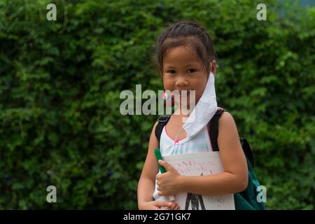 Little asian girl wearing face mask with green back pack holding stationery in green nature  park background. Back to school concept. Stock Photo