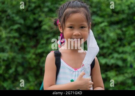 Little asian girl wearing face mask with green back pack holding stationery in green nature  park background. Back to school concept. Stock Photo