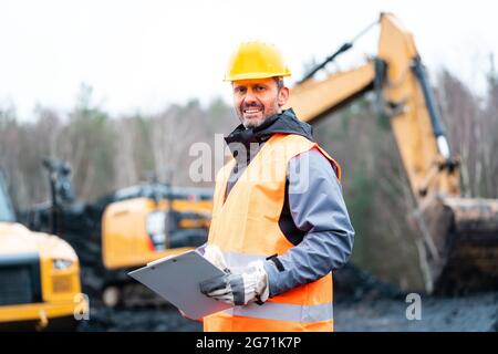 Portrait of a quarry worker standing in front of excavator smiling Stock Photo