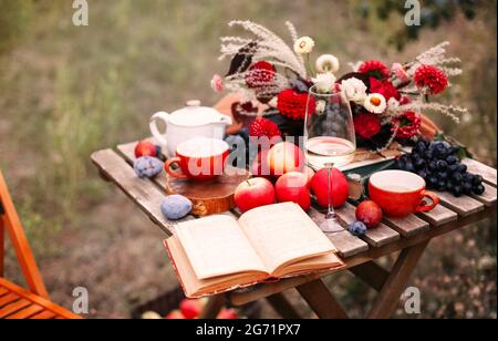 Opened book and glass of wine placed near dishware and bouquet of fresh flowers amidst assorted fruits on wooden table on autumn day in garden Stock Photo