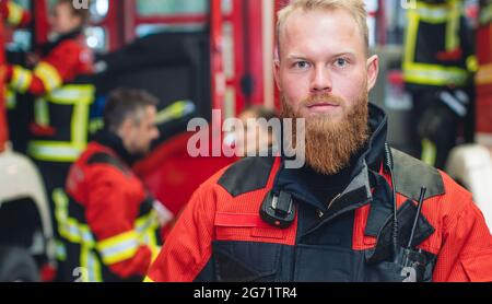 Young and determined fire fighter holding his helmet, with team working in the background Stock Photo