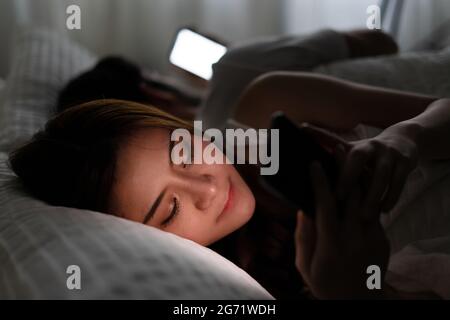 Young couple conflict in bed. happy smiling woman turned her back to man, reading message on mobile phone, trying to peek at screen. Cheating and infi Stock Photo