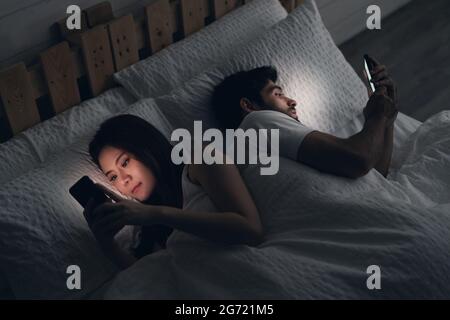 Young couple conflict in bed. happy smiling woman turned her back to man, reading message on mobile phone, trying to peek at screen. Cheating and infi Stock Photo